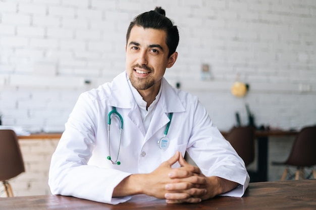 Портрет дружелюбного врача-терапевта-мужчины в белой форме со стетоскопом, сидящего за столом в больничном кабинете и счастливо смотрящего в камеру