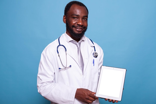 Photo portrait of friendly medic in lab coat with stethoscope presenting digital tochscreen computer with mockup display on blue background. smiling medical doctor in hospital uniform holding white screen d