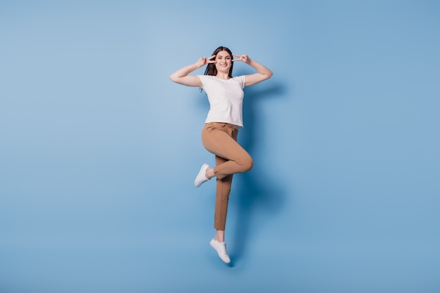 Portrait of friendly positive active lady jump v-signs cover eyes posing on blue background