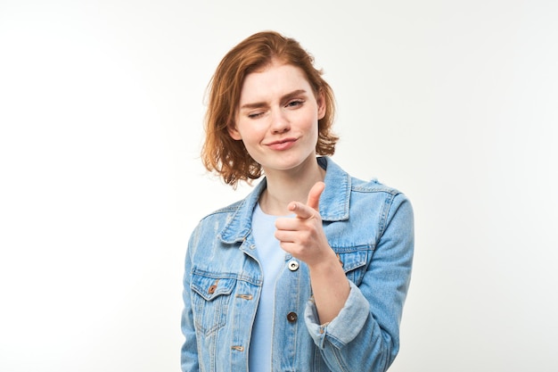 Portrait of friendly redhead girl chooses you points finger at the camera isolated on white background