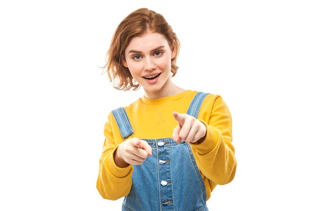 Portrait of friendly redhead girl chooses you points finger at the camera isolated on white background