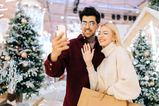 Portrait of friendly young couple talking looking at smartphone\
having video call giving online new year greetings standing in hall\
of celebrate shopping mall