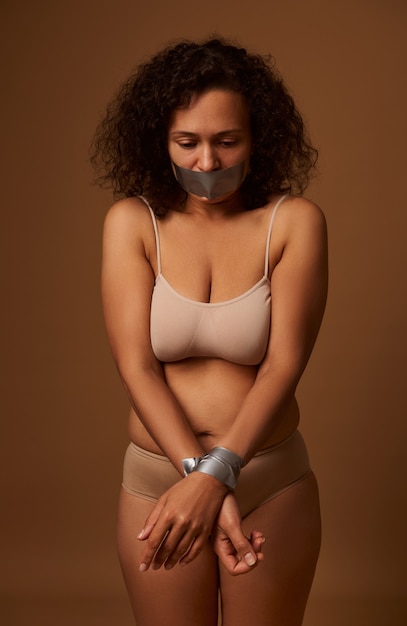 Portrait of frightened desperate mixed race Hispanic woman with sealed mouth looking down with hopelessness, isolated on dark beige background. International Day to Eliminate Violence against Women.