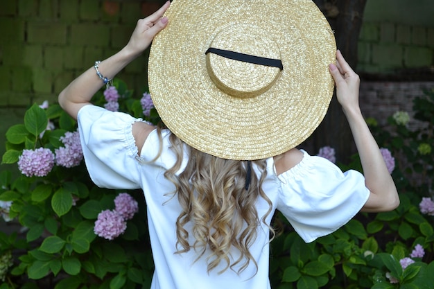 Portrait from back of slim stunning woman looking at pink roses Attractive girl in stylish hat posing in front of flowers