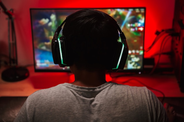 Portrait from back of teenage gamer boy looking at screen and playing video games on computer in dark room wearing headphones