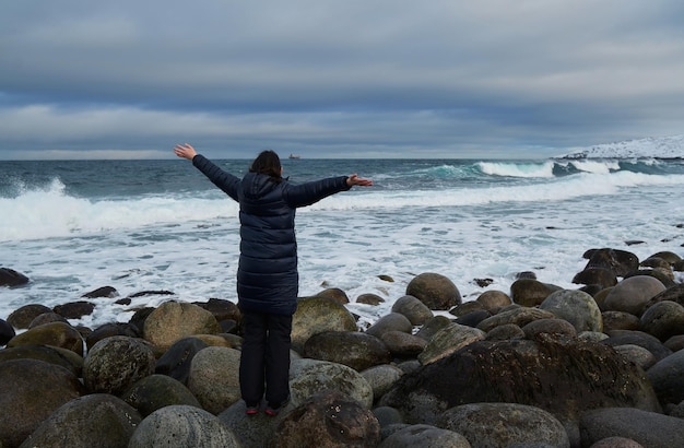 Portrait from back of warmly dressed woman in defocus standing in front of sea looking far away looking at winter rough water in slow motion