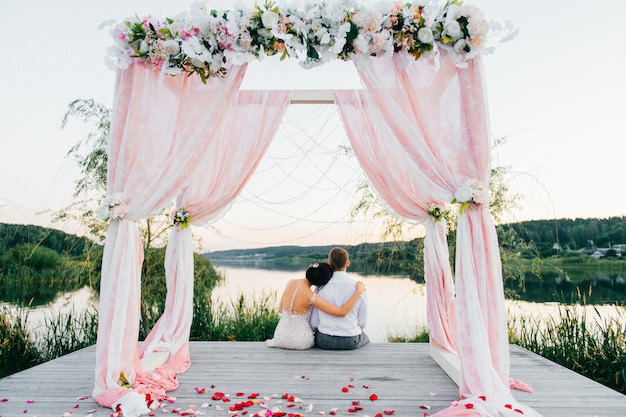 Photo portrait from behind of a romantic wedding couple siiting ar wedding arch and looking at beautiful landscape.