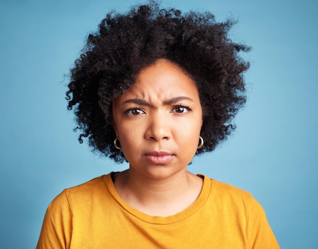 Portrait frown and anger with an african woman on a blue background in studio looking mad or serious face bad or negative and an unhappy young female person with an afro feeling frustrated