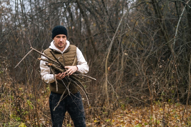 Portrait of frozen tourist male wearing warm clothes collecting dry deadwood for fire in forest on overcast cold day