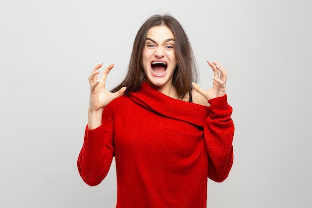 Портрет расстроенной и злой женщины, громко кричащей в красном свитере на светло-сером фоне