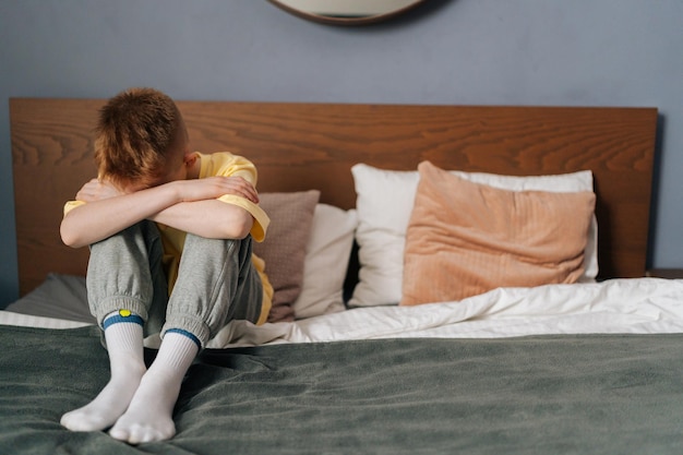 Портрет расстроенного маленького мальчика, обнимающего колено, рыдающего с опущенной головой и плачущего, сидящего в одиночестве на кровати в спальне Несчастный депрессивный маленький ребенок отдыхает в одиночестве