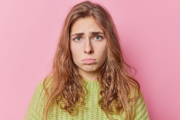 欲求不満の気分を害した若いきれいな女性の肖像画は、カメラを悲しそうに見ている唇を見て気分を害している何かが屋内で緑のニットセーターのポーズを着ていることについて不平を言うことに悩まされている