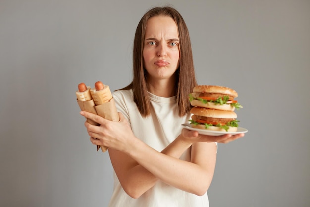 회색 배경 위에 격리된 흰색 티셔츠를 입은 핫도그와 큰 샌드위치를 들고 있는 좌절한 여성의 초상화는 패스트푸드를 먹는 것이 건강에 매우 해롭다고 생각하며 손을 교차시킵니다.