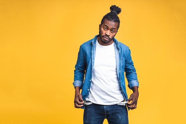 Foto ritratto dell'uomo di colore afroamericano preoccupato frustrato con la barba in tasche vuote casuali che mostrano che non ho gesto dei soldi, bancarotta. isolato su sfondo giallo.