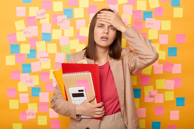 Portrait of frustrated young adult woman having terrible headache or high temperature studies hard holds diary with papers posing against yellow wall with colorful sticky post notes