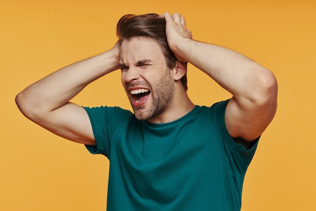 Portrait of frustrated young man holding head in hands and making a face while standing against yellow wall