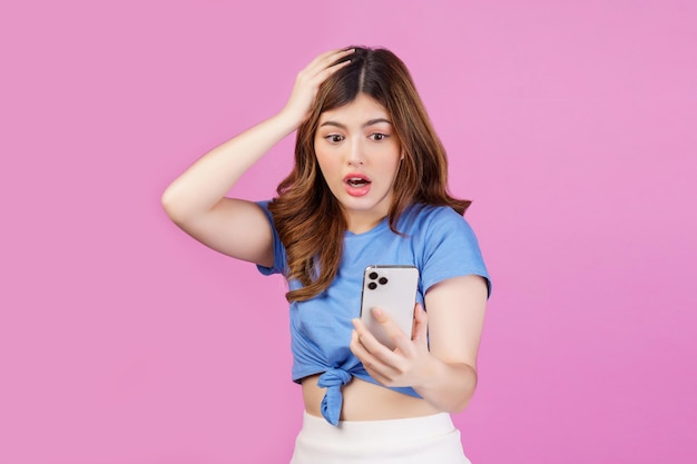 Photo portrait of frustrated young woman using mobile phone in her hands while standing isolated over pink background stressed with hand on head