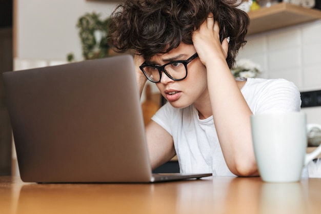 Портрет разочарованной молодой женщины в очках, работающей на портативном компьютере дома утром