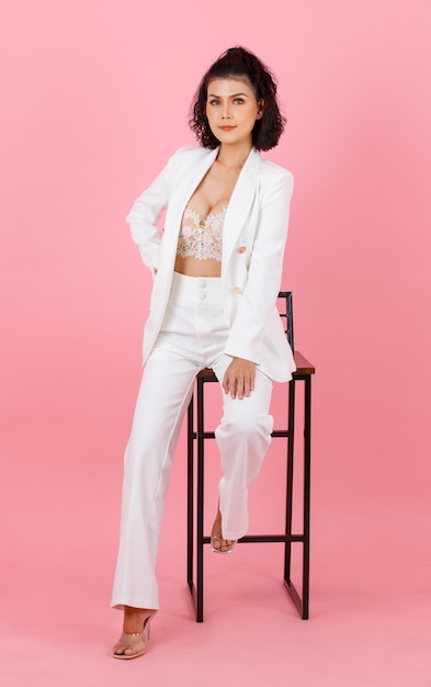 Portrait full body studio shot of Asian sexy curly hairstyle successful businesswoman in white fashionable casual suit with lace lingerie crop top bra sitting on tall chair posing on pink background