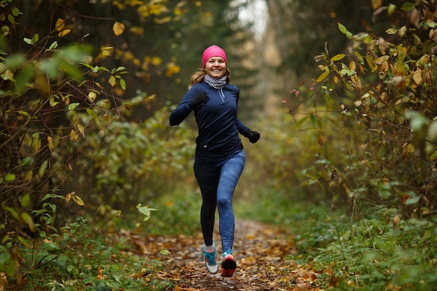 Portrait in full growth of sporty young woman in training suit jogging in colorful forest. Happy model woman in motion