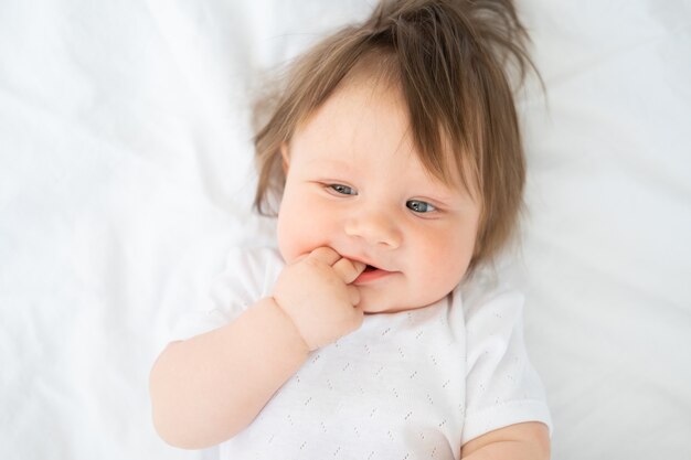 Portrait of funny baby boy with finger in a mouth smiling and lying on a white bedding at home.