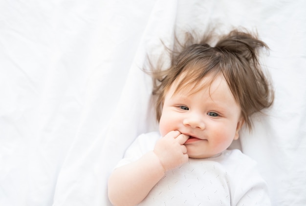 Portrait of funny baby girl with finger in a mouth smiling and lying on a white bedding at home
