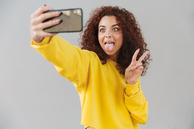 Il ritratto di bella donna riccia divertente isolata sopra la parete grigia facendo uso del telefono cellulare prende un selfie con il gesto di pace che mostra la lingua.