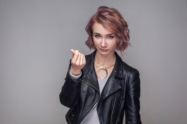 Portrait of funny beautiful girl with short hairstyle and makeup in casual style black leather jacket standing with money gesture and looking at camera. indoor studio shot, isolated on grey background