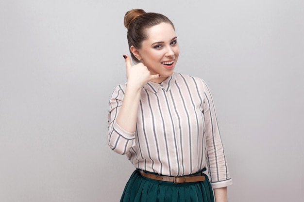 Portrait of funny beautiful young woman in striped shirt with makeup and collected ban hairstyle, standing with call gesture and looking at camera. indoor studio shot, isolated on grey background.