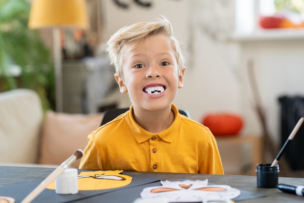 portrait of funny boy with fangs like vampire sitting at table and making decorations while preparing for halloween