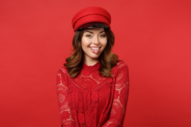 Portrait of funny cheerful young woman in lace dress, cap looking camera showing tongue isolated on bright red wall background in studio. People sincere emotions lifestyle concept. Mock up copy space.