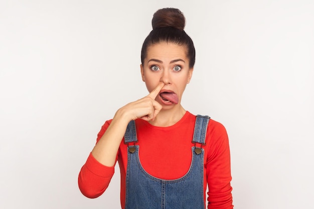 Portrait of funny comical girl with hair bun in denim overalls picking nose and looking with surprised goofy face, showing tongue out, removing boogers. indoor studio shot isolated on white background