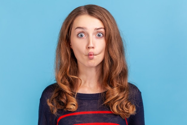 Portrait of funny crazy woman standing and looking with crossed\
eyes and fish lips, having fun, humor, wearing striped casual style\
sweater. indoor studio shot isolated on blue background.