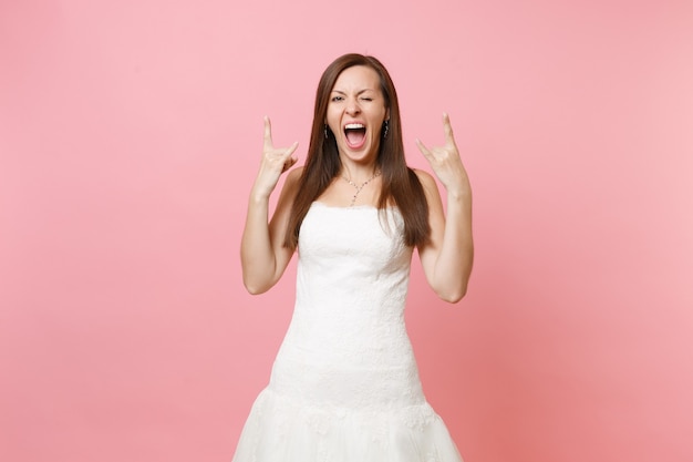 Portrait of funny crazy woman in white dress standing blinking and showing rock-n-roll sign