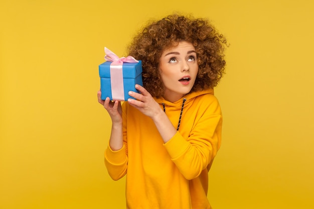 Portrait of funny curious curlyhaired woman in urban style hoodie holding present box near ear and listening what39s inside in anticipation of dream gift studio shot isolated on yellow background