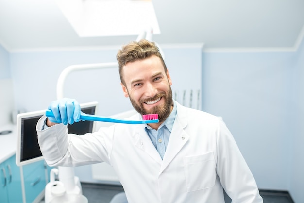 Портрет забавного дантиста в униформе с большой зубной щеткой в стоматологическом кабинете