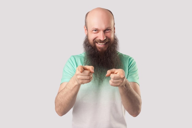 Portrait of funny excited middle aged bald man with long beard, green t-shirt standing, pointing and looking at camera with toothy smile and happiness. indoor studio shot isolated on grey background