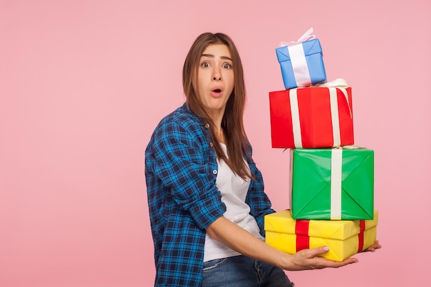 たくさんの重いプレゼントボックスを持って、たくさんの誕生日プレゼントに驚いた、驚いたコミカルな表情でカメラを見ている面白い女の子の肖像画。ピンクの背景に分離された屋内スタジオショット