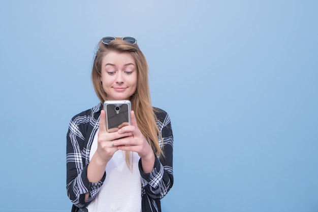 Портрет смешной девушки используя smartphone на голубой предпосылке.
