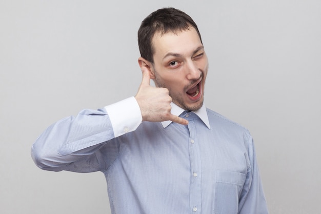 Portrait of funny handsome bristle businessman in classic light blue shirt standing with call gesture and looking at camera winking. indoor studio shot, isolated on grey background copyspace.