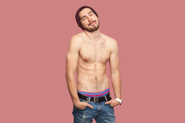 Portrait of funny handsome naked shirtless young bearded man standing, hands on pocket, looking at camera, tongue out. flirting and playful sexy face. indoor studio shot, isolated on pink background.