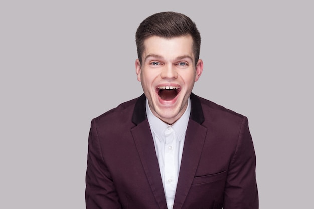 Portrait of funny handsome young man in violet suit and white shirt, standing, looking at camera with open mouth and laughing. indoor studio shot, isolated on grey background.