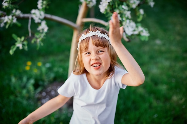 Portrait of funny kid girl 5-6 year old holding flower standing in blossom spring garden, Wearing white dress and floral wreath outdoors, Spring season is coming.