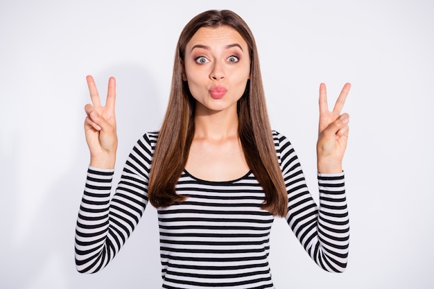 Portrait of funny lady sending air kisses making v-signs wearing striped sweater isolated over white wall