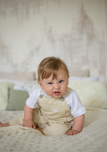 Portrait of a funny little boy in a beige jumpsuit sitting on a bed in a room and making grimaces