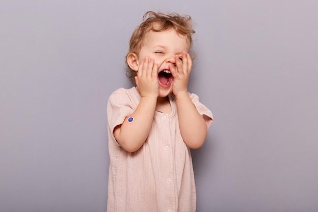 Portrait of funny little caucasian girl posing isolated over gray background infant baby with blond wavy hair laughing with open mouth having fun touching face