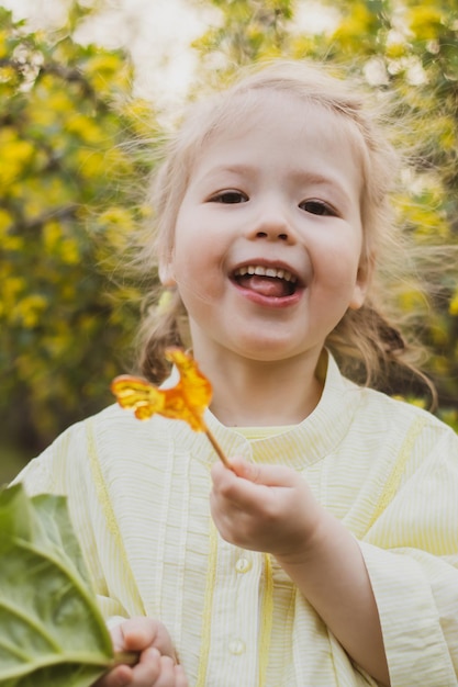 Portrait of funny little girl in yellow dress sucking candy on the background flowering garden