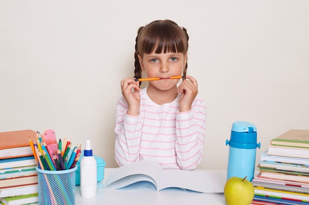 Portrait of funny little schoolgirl with pigtails wearing striped shirt sitting at the desk surrounded with school supplies writing in exercises holding pen in mouth