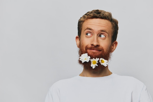 Портрет забавного человека в белой футболке с цветами ромашками в бороде на белом изолированном фоне копирует место Концепция праздника и поздравления