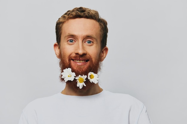 Photo portrait of a funny man in a white tshirt with flowers daisies in his beard on a white isolated background copy place holiday concept and congratulations